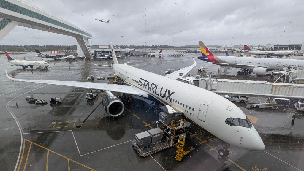 A Starlux Airlines plane at the airport terminal, with luggage being loaded and other aircraft visible in the background under overcast skies.